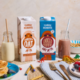 Three Robins Barista and Choccoriffic oat milk cartons beside glass of oat milk with straw and glass of chocolate milk with straw. Surrounded by kids toy animals, crayons, plate of fruit, plate of pancakes, bowl of cereal