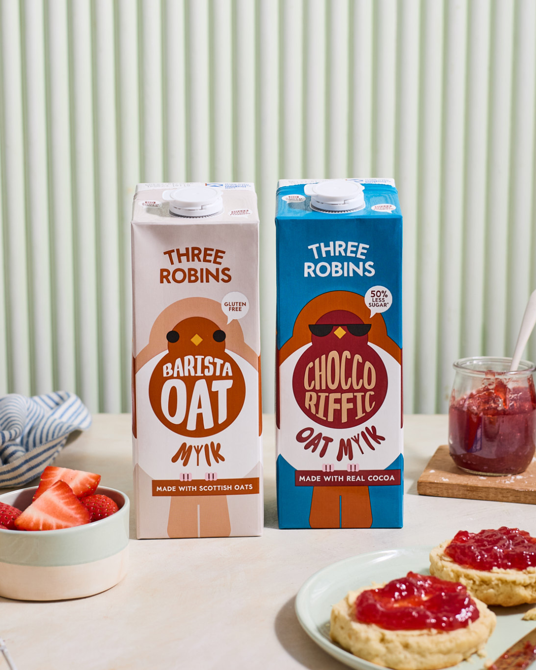 Three Robins Barista and Choccoriffic oat milk cartons beside bowl of cut strawberries and scone with jam