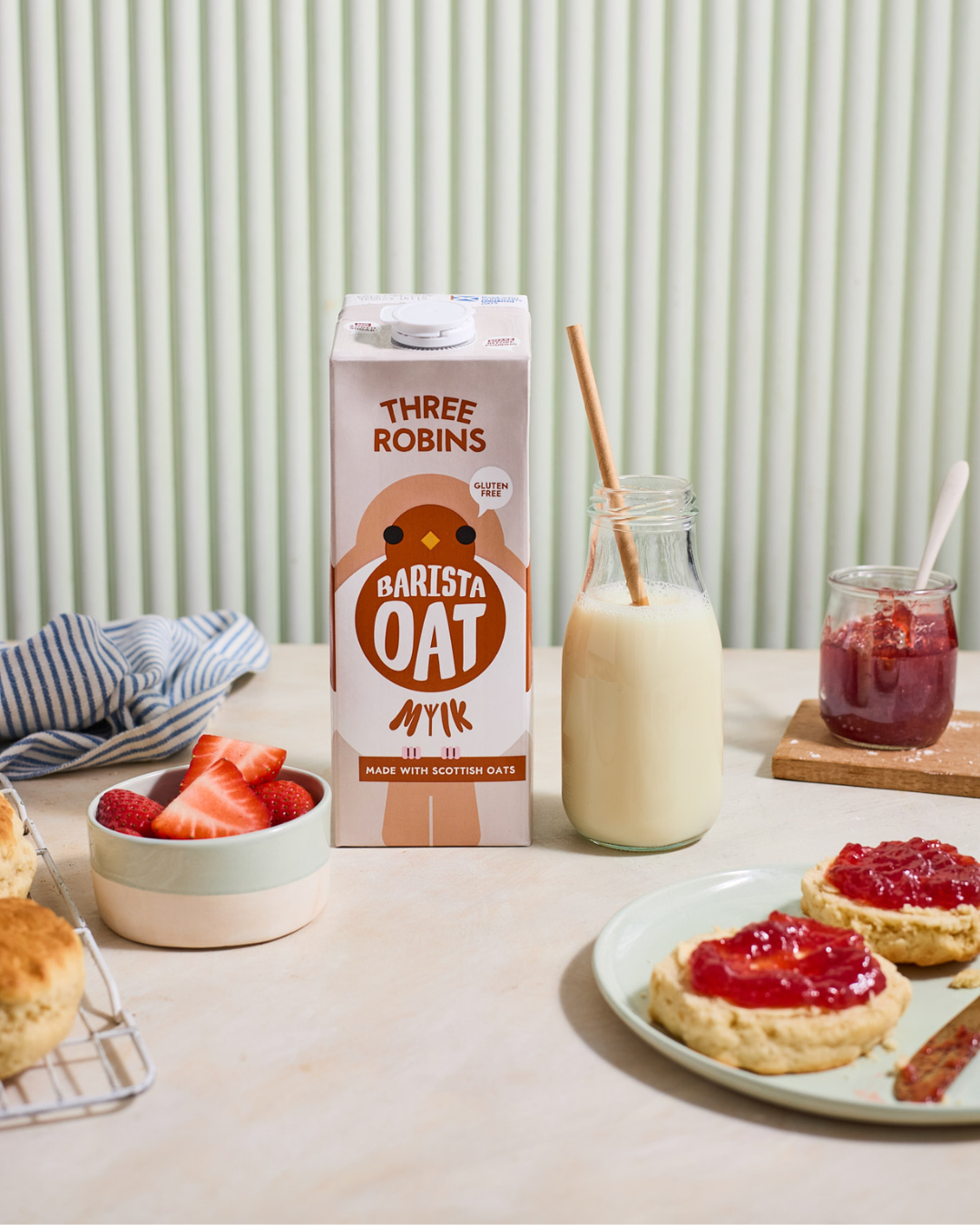 Three Robins Barista oat milk on table with strawberries, glass of oat milk with straw, scone with jam and jam jar