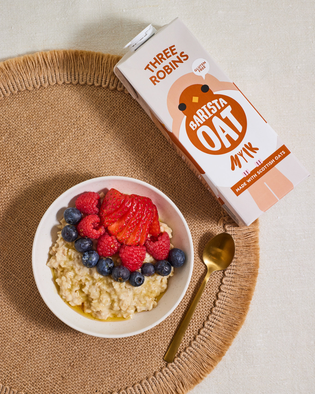 Three Robins Barista oat milk carton laying flat beside bowl of porridge with fruit and golden syrup on top
