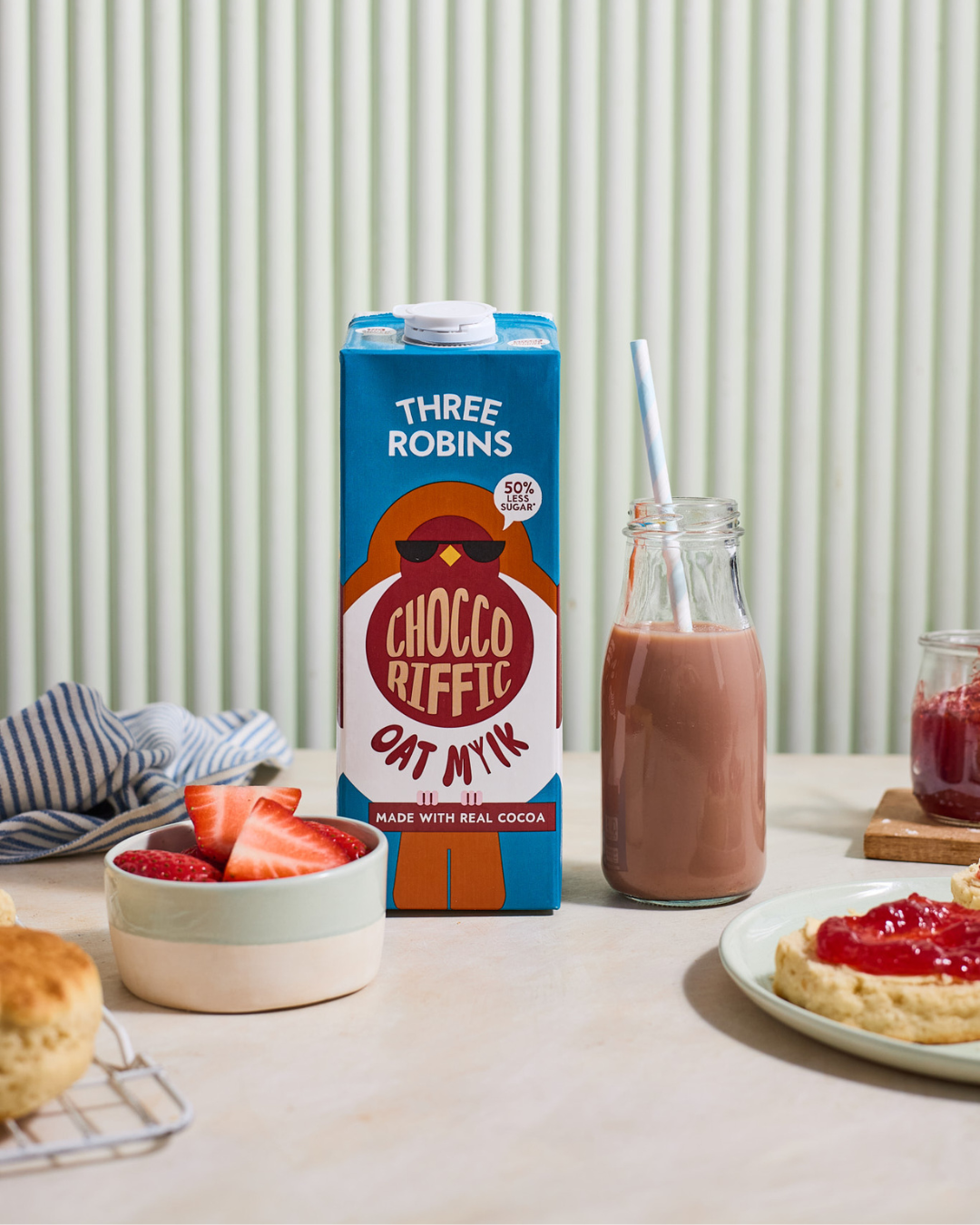 Three Robins Choccoriffic oat milk carton beside bowl of cut strawberries, glass of chocolate oat milk with straw and scone with jam