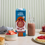 Three Robins Choccoriffic oat milk carton beside bowl of cut strawberries, glass of chocolate oat milk with straw and scone with jam