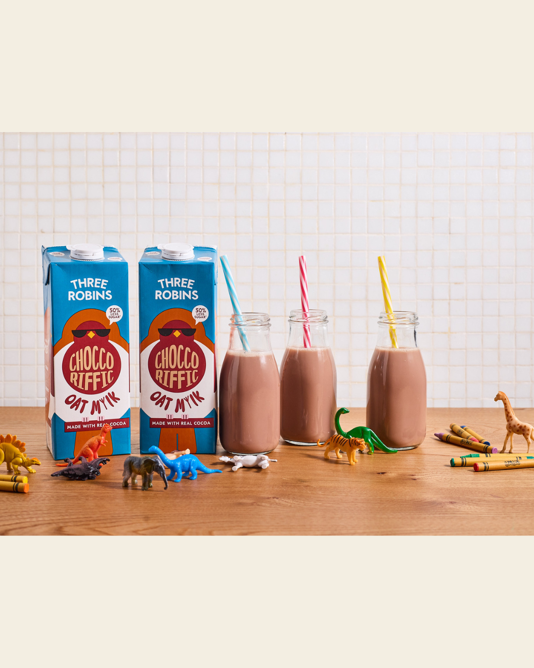 Three Robins Choccoriffic oat milk cartons beside glasses of chocolate oat milk with straw and kids toy animals and crayons
