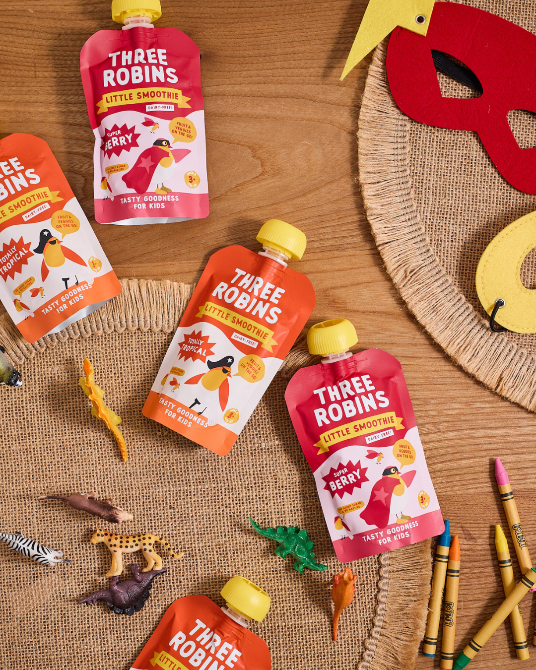 Three Robins Little Smoothie pouches on table surrounded by kids toy animals and dinosaurs, crayons and felt masks.