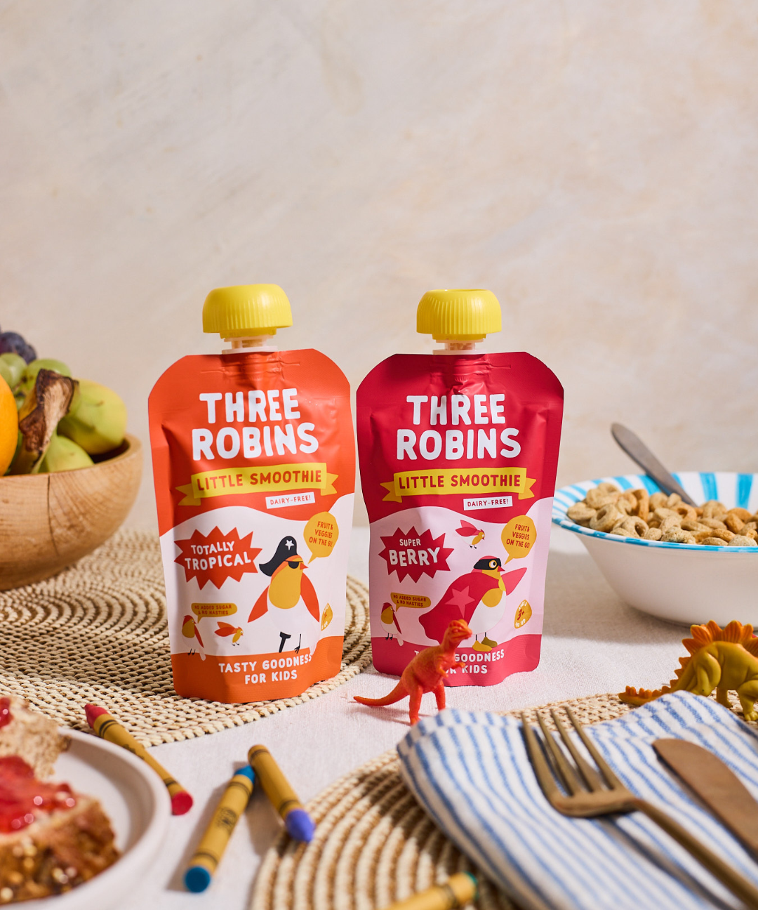 Three Robins Totally Tropical Little Smoothie pouches side by side on family breakfast table surrounded by crayons, toy dinosaurs and bowls of cereal and fruit