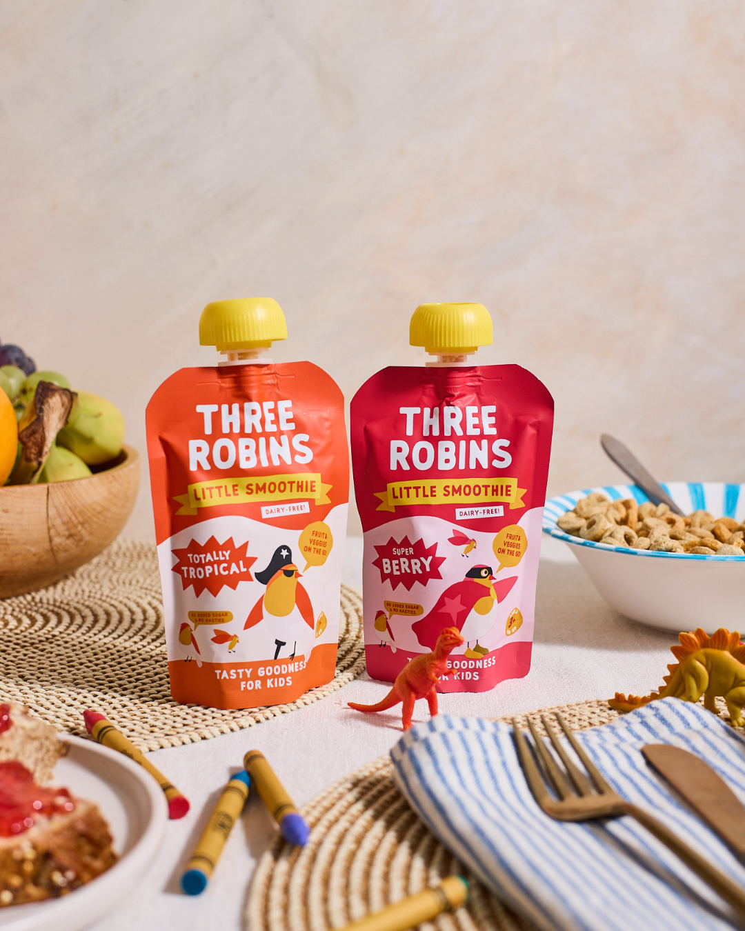 Three Robins Totally Tropical Little Smoothie pouches side by side on family breakfast table surrounded by crayons, toy dinosaurs and bowls of cereal and fruit