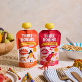 Three Robins Totally Tropical Little Smoothie pouches side by side on family breakfast table surrounded by crayons, toy dinosaurs and bowls of cereal and fruit