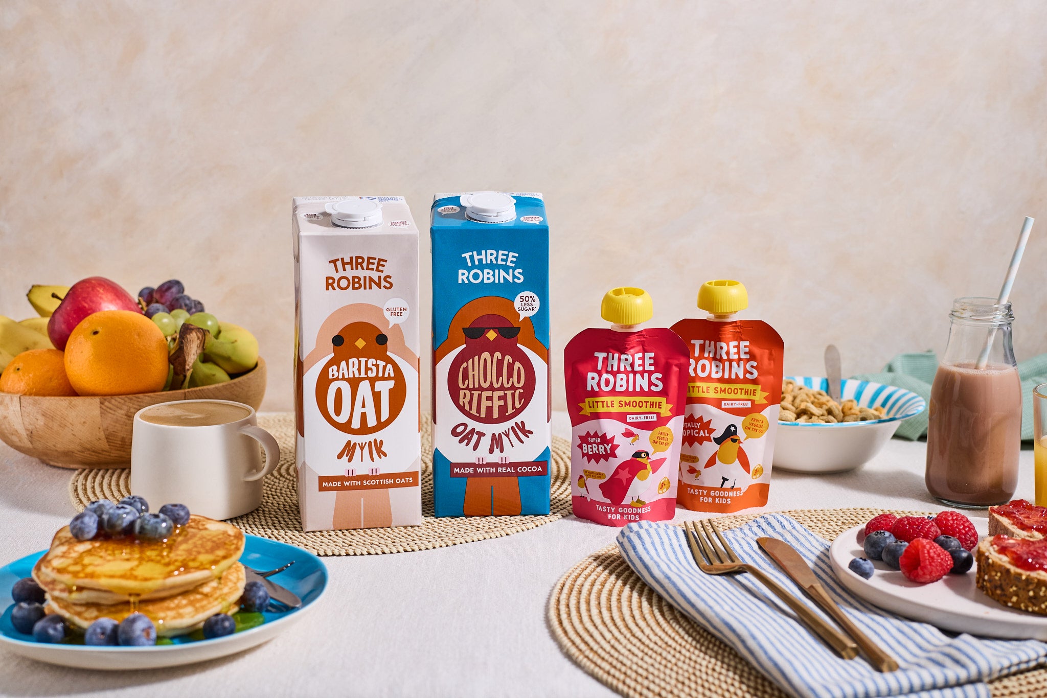 Three Robins full range on a family breakfast table. Barista oat milk carton beside choccoriffic oat milk carton and the super berry and totally tropical pouches. In the background there is a mug of coffee, a glass with chocolate oat milk and a straw, a bowl of cereal, a plate of pancakes and toast with jam and fruit