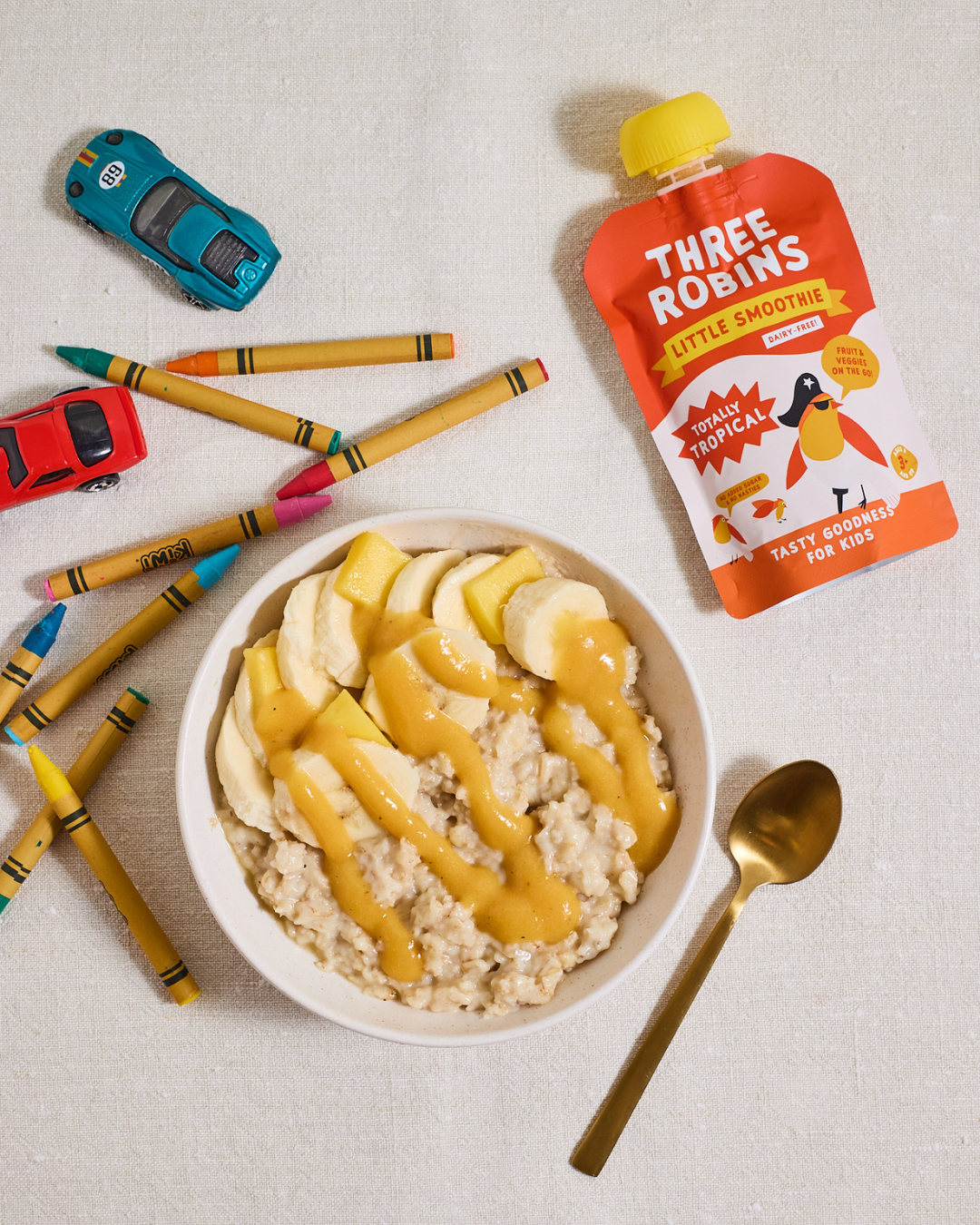 Three Robins Totally Tropical Little Smoothie pouch lying beside bowl of porridge with smoothie drizzled on top, kids crayons and toy cars