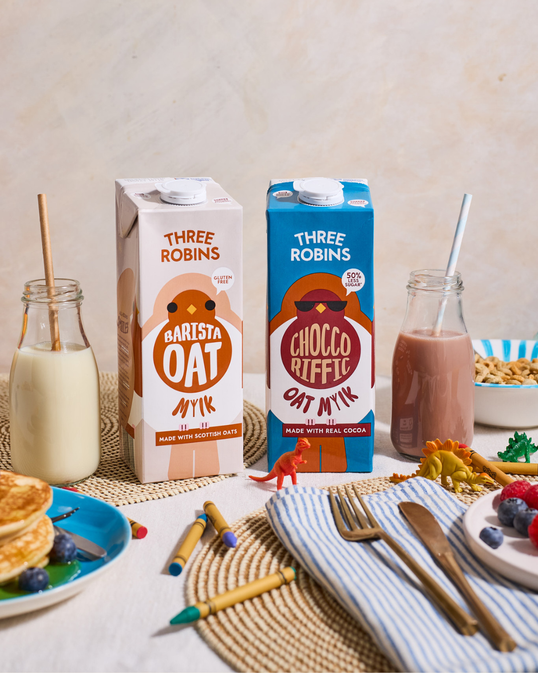 Three Robins Barista and Choccoriffic oat milk cartons beside glass of oat milk with straw and glass of chocolate milk with straw. Surrounded by kids toy animals, crayons, plate of fruit, plate of pancakes, bowl of cereal