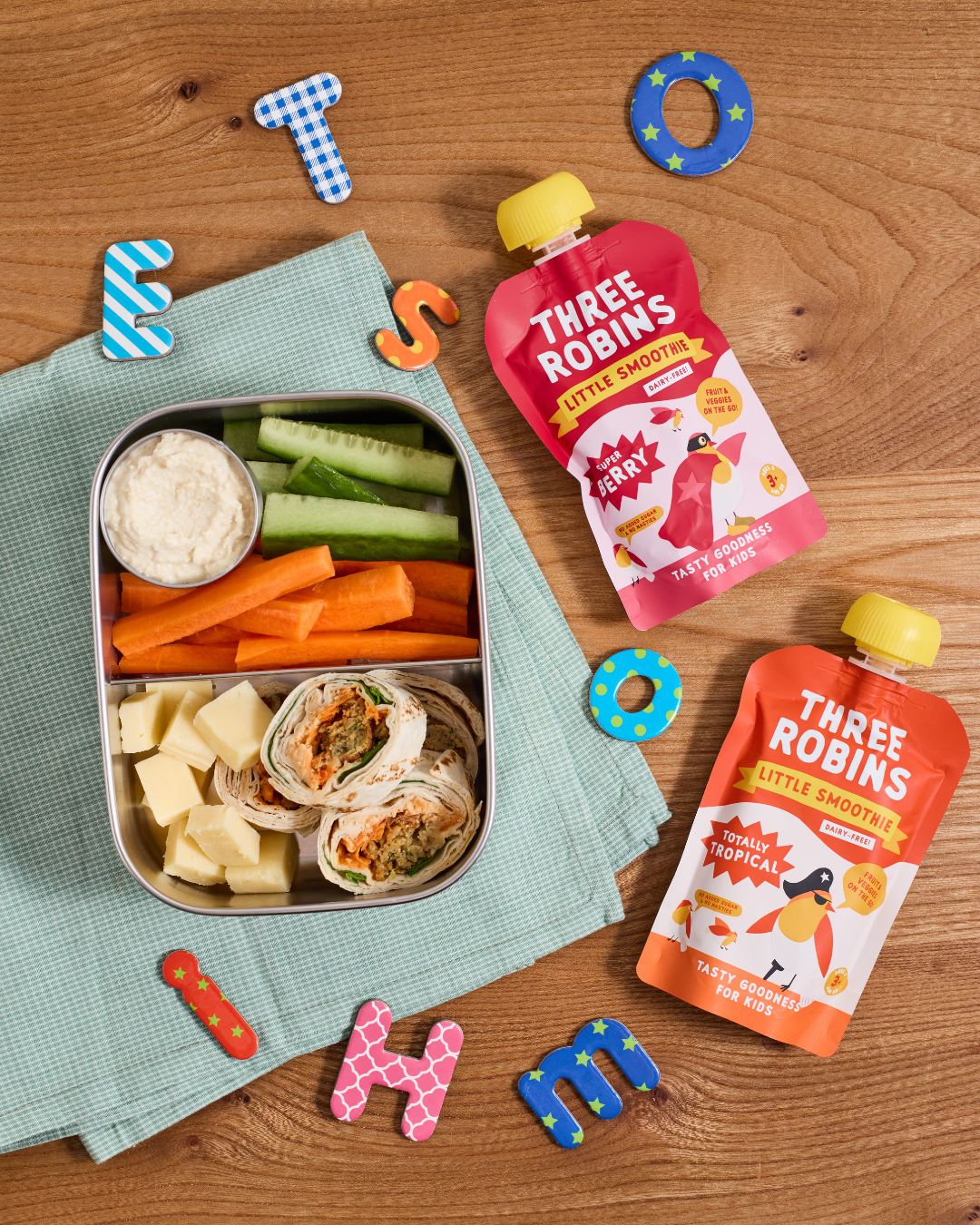 Three Robins Super Berry and Totally Tropical Little Smoothie pouches beside kids spelling letters and kids lunch box filled with snacks