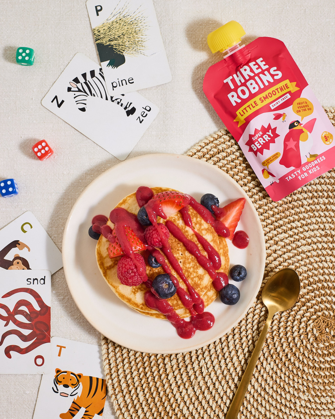 Three Robins Super Berry Little Smoothie pouch beside pancakes with smoothie drizzled on top with fresh fruit and kids animal cards and coloured dice on table