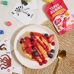 Three Robins Super Berry Little Smoothie pouch beside pancakes with smoothie drizzled on top with fresh fruit and kids animal cards and coloured dice on table