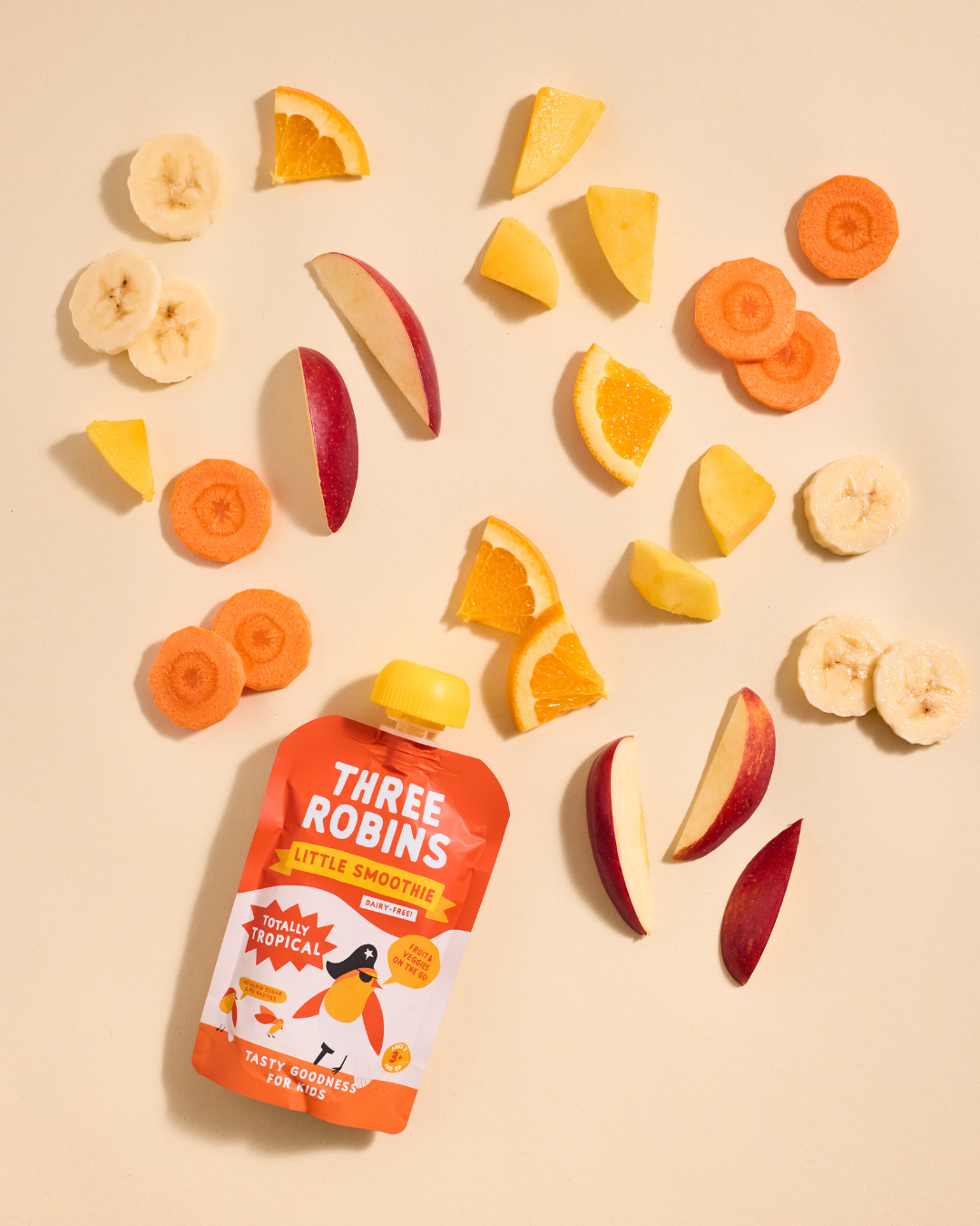 Three Robins Totally Tropical Little Smoothie pouch surrounded by cut fruit and veg including carrots, apples, bananas, oranges and mangoes