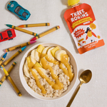 Three Robins Totally Tropical Little Smoothie pouch lying beside bowl of porridge with smoothie drizzled on top, kids crayons and toy cars